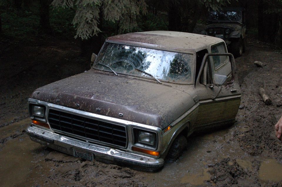Mud-covered Ford Bronco stuck in deep mud on a forest trail, driver door open.