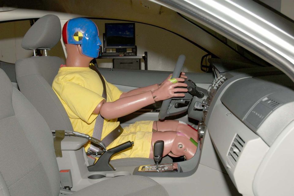 Crash test dummy in driver's seat, wearing yellow shirt and blue helmet. Car interior visible