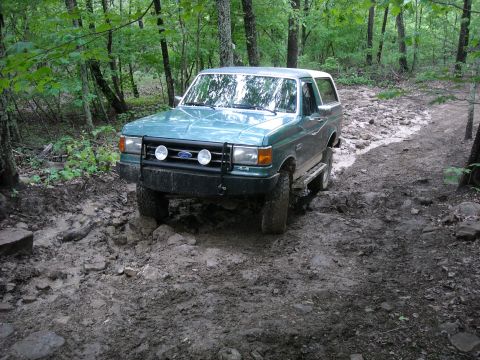 1979 Ford F-150 - A teal Ford Bronco navigates a challenging muddy and rocky off-road path through a forest.