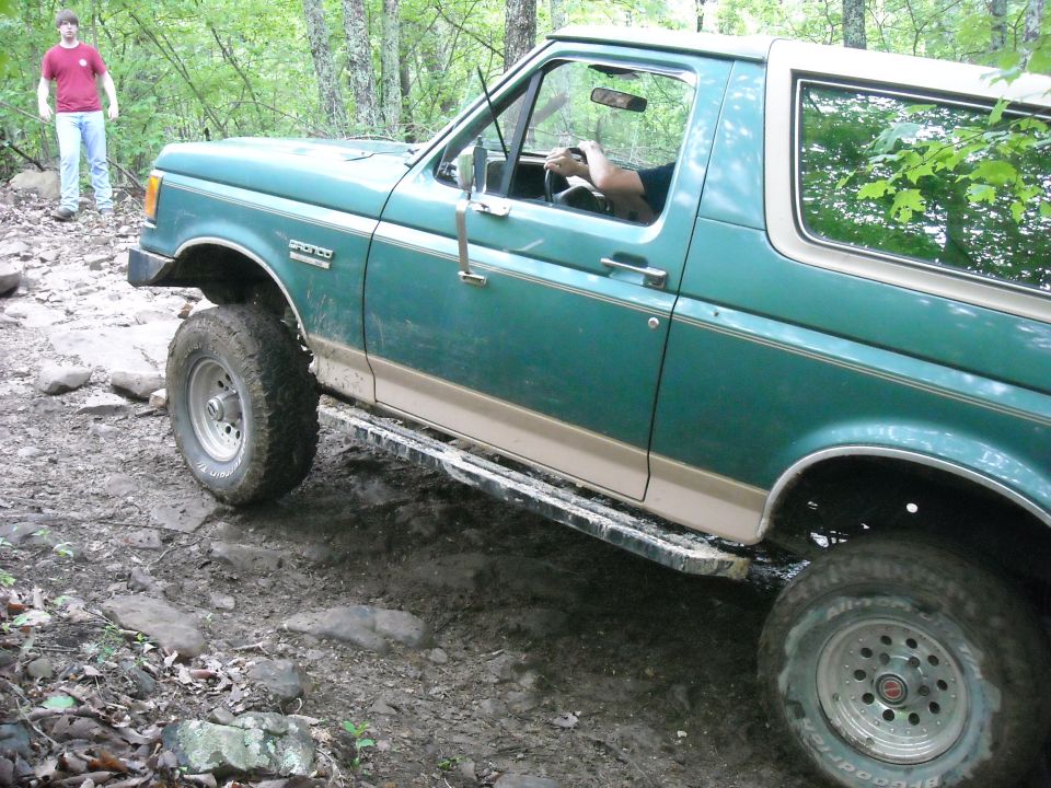 Green Ford Bronco with tan trim driving on a rocky trail, person in driver's seat, another person standing in background