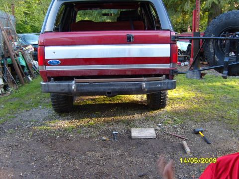 1989 Ford Bronco - Rear view of a red Ford Bronco with a silver stripe and an aftermarket bumper, showing a spare tire carrier on the side.