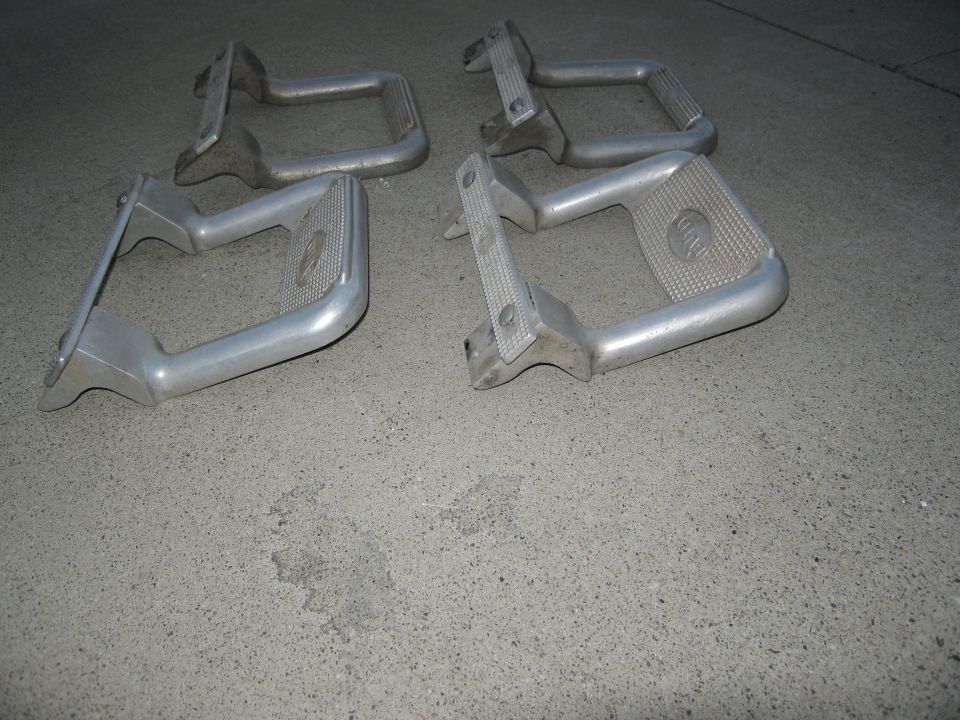 Four aluminum running board steps with textured grip pads and visible mounting brackets on a concrete surface
