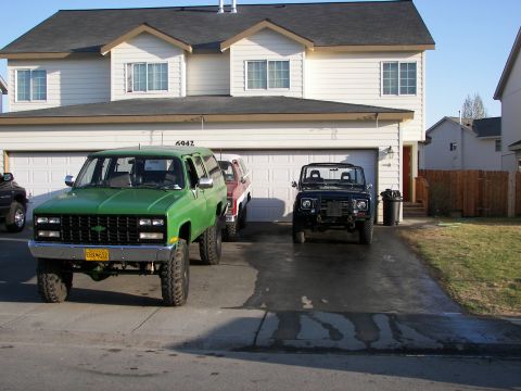 1990 Chevrolet Suburban - Two SUVs, a green Chevrolet Suburban and a black Jeep, are parked in a driveway in front of a house.
