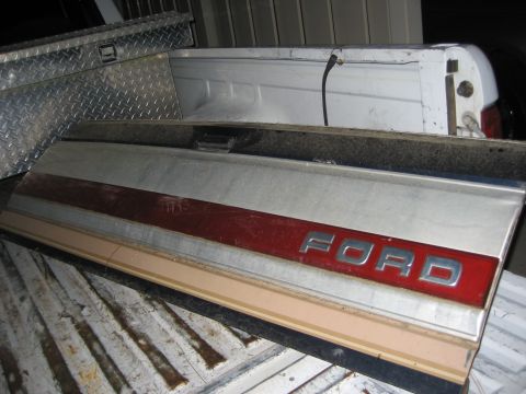 1983 Ford F-250 - A Ford tailgate panel with a red stripe and chrome lettering is stored in the bed of a pickup truck.