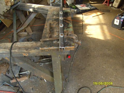 1989 Ford Bronco - A metal bar with circular cutouts is secured to a workbench in a cluttered workshop setting.