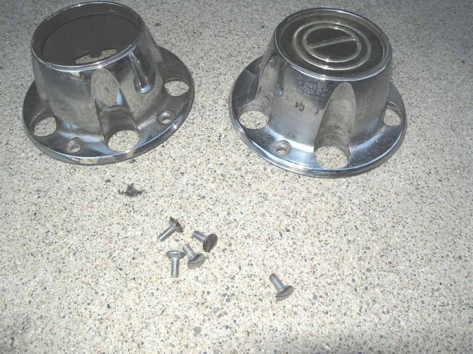 Two chrome wheel hubcaps and four screws on a concrete surface