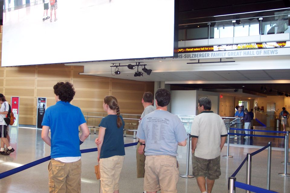 People stand in a lobby looking at a large screen and a news ticker