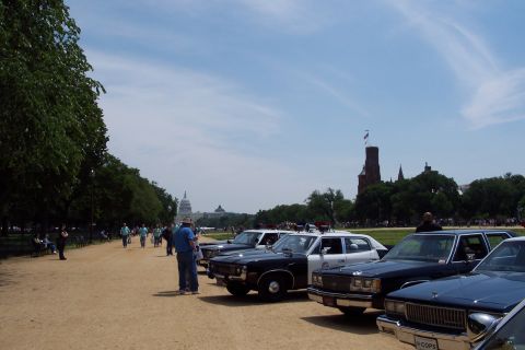 2001 Ford Crown Victoria - Several vintage police cars are lined up on a sunny day, with the Washington Monument visible in the distance.