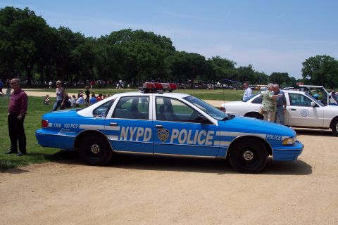 2001 Ford Crown Victoria - A blue and white NYPD police car is parked on a dirt lot with people in the background.
