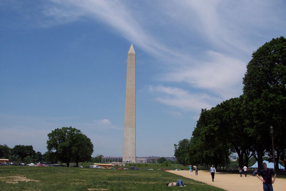 Washington Monument under a blue sky with scattered clouds, with people walking on a path and cars parked in the distance
