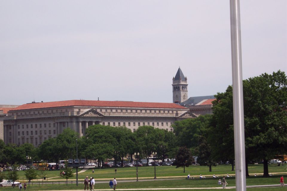Large classical building with a clock tower, surrounded by trees and a park with people