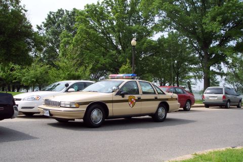 2001 Ford Crown Victoria - A tan police car with a light bar and Maryland insignia is parked next to other vehicles.