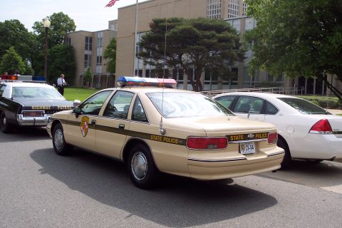 2001 Ford Crown Victoria - A tan state police car is parked behind a black highway patrol vehicle, with a white sedan visible to the right.