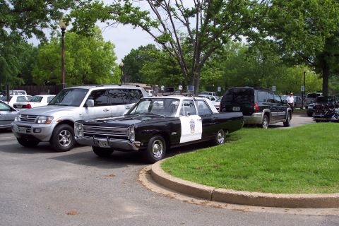 2001 Ford Crown Victoria - A vintage Plymouth police car is parked alongside modern SUVs in a lot.