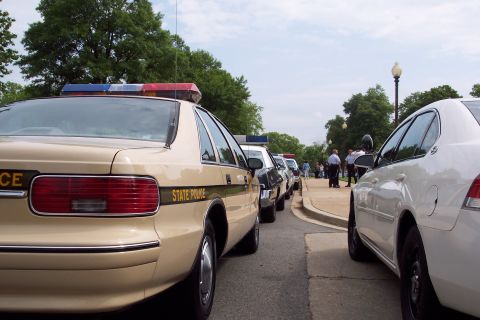 2001 Ford Crown Victoria - A line of tan and white police cars are parked on the side of a road with trees and people in the background.