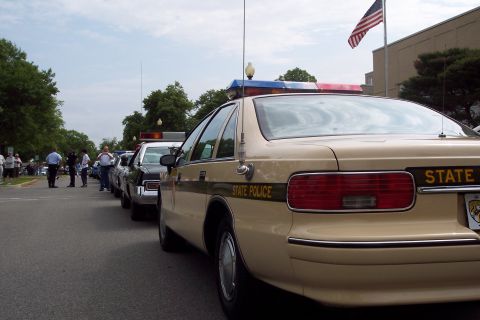 2001 Ford Crown Victoria - A line of tan and black state police cars are parked on a street, with an American flag flying in the background.