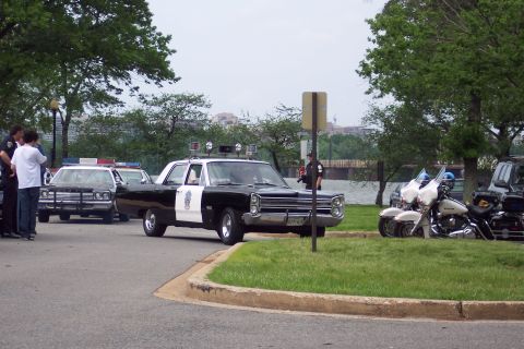 2001 Ford Crown Victoria - A vintage black and white Plymouth police car is parked near a waterfront with other vehicles and people.