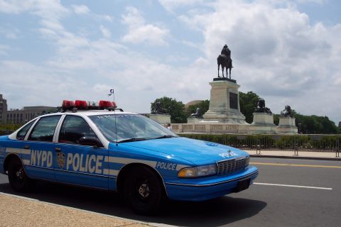 2001 Ford Crown Victoria - A blue and white NYPD police car is parked on a street with a monument in the background under a cloudy sky.