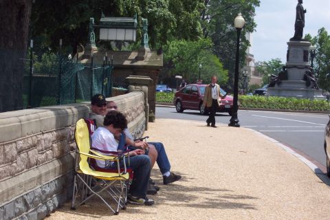 2001 Ford Crown Victoria - A red sedan drives by a group of people sitting on a stone wall on a sunny day.