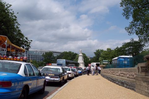 2001 Ford Crown Victoria - A procession of police vehicles and a bus are lined up on a street, with a monument and buildings visible in the distance.