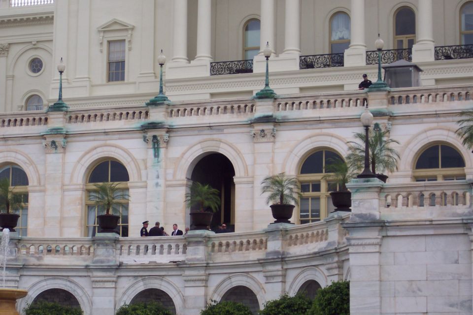 People stand on a balcony of a large white building with arched windows and columns