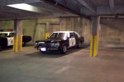 2001 Ford Crown Victoria - Two police cars are parked in a concrete garage. The front car has a push bumper and a light bar on top.