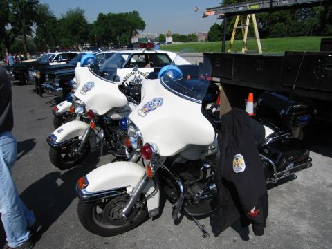 1991 Ford LTD Crown Victoria - Two white police motorcycles with emblem decals are parked together on pavement, a dark uniform jacket draped over the saddlebag of one.