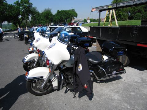 1991 Ford LTD Crown Victoria - line of white police motorcycles with department emblems sits beside a flatbed trailer, with vintage patrol cars parked in the background.