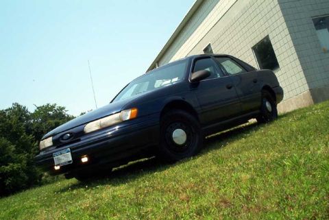 1993 Ford Taurus - Low-angle three-quarter front view of a dark blue Ford Taurus sedan parked on grass beside a beige block building, headlights and fog lamps…