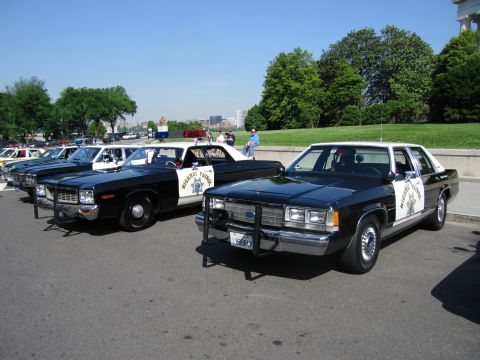 1991 Ford LTD Crown Victoria - black-and-white Dodge and a Ford highway patrol cruiser sit parked at an angle on pavement, with more patrol cars and onlookers behind.