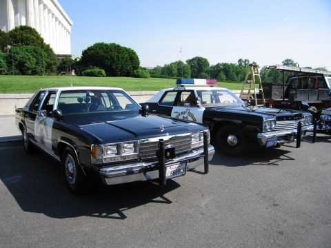 1991 Ford LTD Crown Victoria - Two black-and-white patrol sedans and an armored vehicle are parked in front of a large columned marble building.