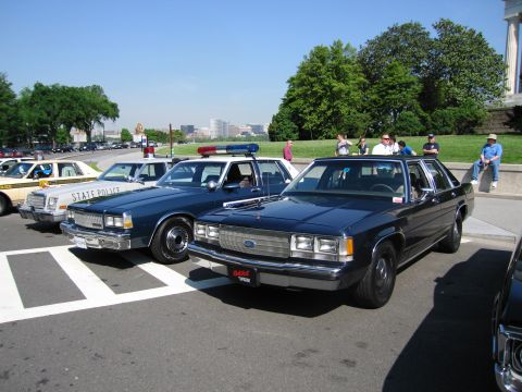 1991 Ford LTD Crown Victoria - dark Ford police sedan is parked beside a STATE POLICE cruiser with a roof lightbar on a sunny day as spectators stand nearby.