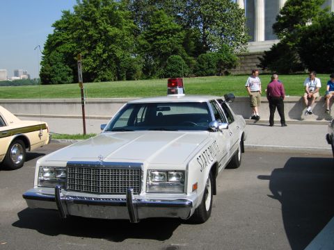 1991 Ford LTD Crown Victoria - white STATE POLICE Chrysler sedan with a chrome grille and red roof beacon is parked on pavement as people sit on a wall behind it.
