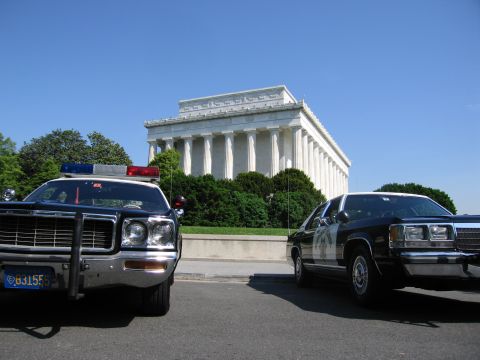 1991 Ford LTD Crown Victoria - front of a patrol car with a roof lightbar and a dark sedan are parked on pavement before the columned Lincoln Memorial.