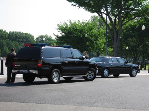 1991 Ford LTD Crown Victoria - black Chevrolet Suburban with roof emergency lights and a black sedan parked curbside near a tree-lined park.