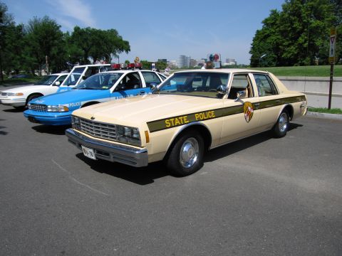 1991 Ford LTD Crown Victoria - tan Chevrolet sedan marked State Police with a Maryland-style crest, lined up beside blue and white police cars.