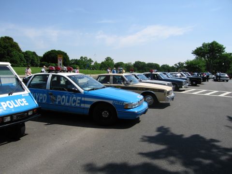 1991 Ford LTD Crown Victoria - diagonal row of vintage and modern police cars, including blue NYPD and tan State Police sedans, parked in a sunny lot.