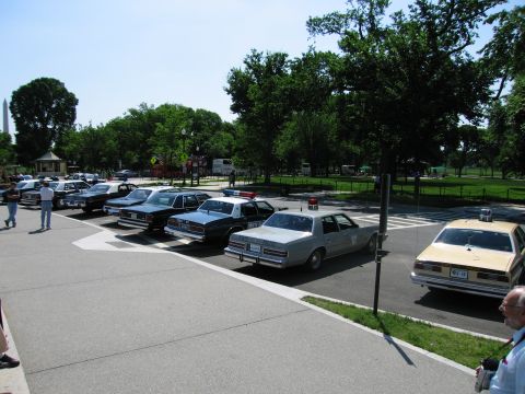 1991 Ford LTD Crown Victoria - Several police sedans, some marked Police, parked in a paved lot with trees and a distant obelisk monument.