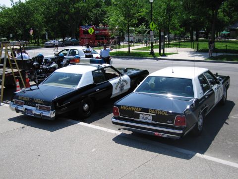 1991 Ford LTD Crown Victoria - Two black-and-white Highway Patrol sedans of different eras parked side by side, with a police motorcycle to the left.