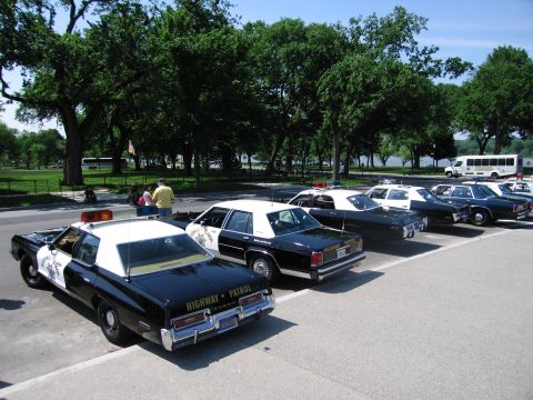 1991 Ford LTD Crown Victoria - group of black-and-white police sedans, one marked Highway Patrol, parked in a tree-shaded lot with a van behind.