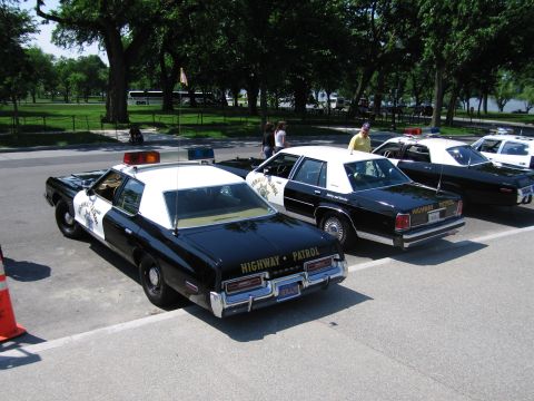1991 Ford LTD Crown Victoria - line of black-and-white Highway Patrol sedans seen from the rear, with people standing alongside under leafy trees.