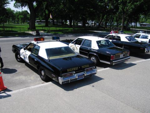 1991 Ford LTD Crown Victoria - Several black-and-white Highway Patrol sedans parked diagonally in a paved lot, with a Canadian flag visible at right.