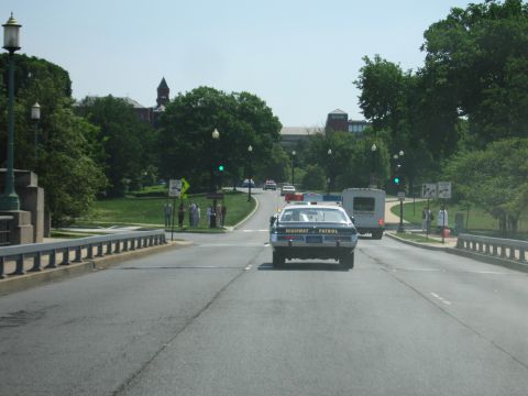 1991 Ford LTD Crown Victoria - blue and white sedan marked Highway Patrol drives away down a wide tree-lined boulevard toward a brick building.