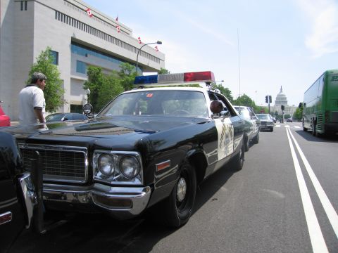 1991 Ford LTD Crown Victoria - black-and-white police sedan with light bar and CHP-style star parked on a city street, with the US Capitol dome in the distance.