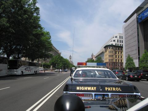1991 Ford LTD Crown Victoria - View over the trunk of a black-and-white Highway Patrol car driving down a city boulevard past a tour bus and buildings.