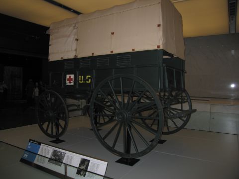 1991 Ford LTD Crown Victoria - dark-green canvas-topped horse-drawn military supply wagon marked U.S. with a red cross, displayed indoors on a museum platform.