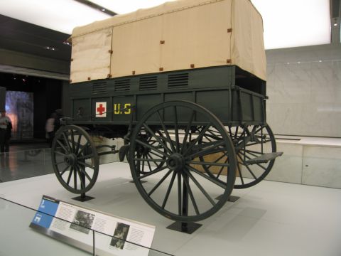 1991 Ford LTD Crown Victoria - green wooden horse-drawn military ambulance wagon with a tan canvas cover and a red-cross U.S. marking sits on a museum display platform.
