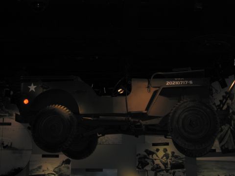 1991 Ford LTD Crown Victoria - olive-drab military jeep with a white star and serial markings hangs suspended in a dimly lit museum exhibit.