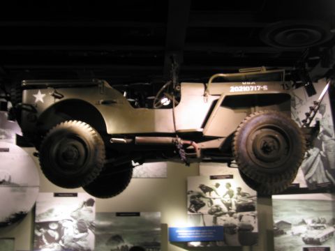 1991 Ford LTD Crown Victoria - khaki military jeep marked USA 20210717-S is mounted high in a dim museum exhibit above wartime photo panels.