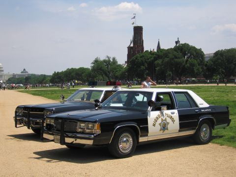 1991 Ford LTD Crown Victoria - Two black-and-white Highway Patrol Ford sedans with push bars sit on the National Mall near the Smithsonian Castle and the Capitol.